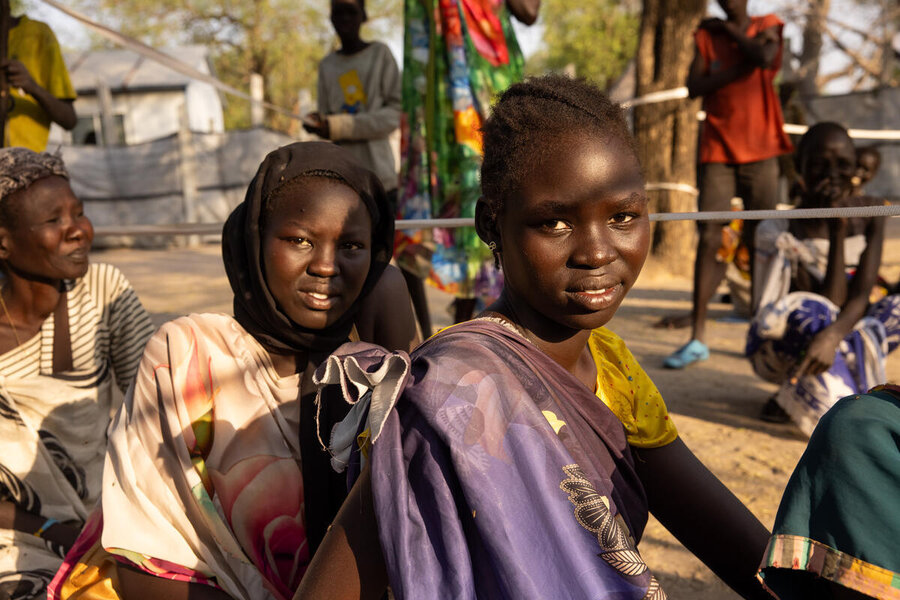 Three girls look into the camera with gentle smiles as they wait sat on the ground a food aid registration in south sudan